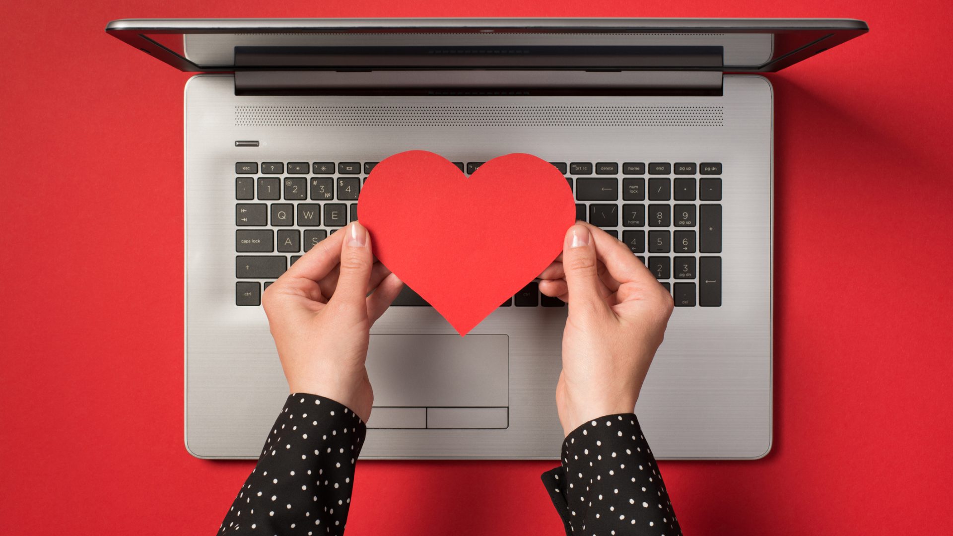 Above photo of grey laptop and hands holding a red paper heart isolated on the red backdrop